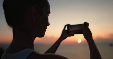 Kadın silueti Tayland 'da dinlenirken telefonuyla gün batımının güzel bir fotoğrafını çekiyor. Kız parlak renkli gökyüzünün tadını çıkarıyor. Açık hava yaşam tarzı, yaz tatili, Kapat.