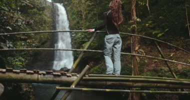 Yürüyüşçü kız Bali adasında orman şelalesinin tadını çıkarıyor. Uzun saçlı turist kadın bambu köprüsünde yürüyor ve güçlü şelalenin keyfini çıkarıyor. Seyahat tatili. Eriyen Şelaleler. Yavaş