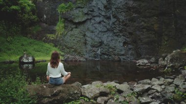 Vahşi bir tropikal ormanda küçük bir şelalenin yanında yoga yapan bir kadın. Huzurlu bir farkındalık ve doğayla bağlantı anının tadını çıkarıyor.