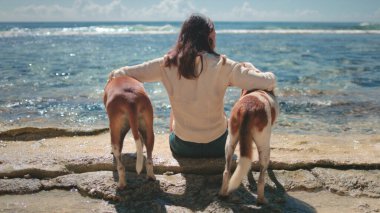 Genç kadın deniz kenarındaki kayalıklarda iki köpeği ile oturuyor, onları kucaklıyor, huzur ve sükunetin tadını çıkarıyor.