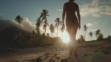 Fransız Polinezyası 'nın yemyeşil palmiye ağaçlarına çerçevelenmiş, gün batımı kumsalında yürüyen bir kadın. Gökyüzü turuncu ve pembenin canlı bir karışımıdır. Tropikal araziye sıcak bir parıltı yayar..