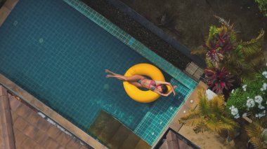 Genç kadın güneşli bir günün tadını çıkarıyor, sarı şişme bir yüzükte lüks bir yüzme havuzunda süzülüyor, yemyeşil tropikal bitki örtüsüyle çevrili.