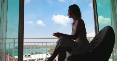 Beyaz kadın silueti balkonda kahve içiyor ve okyanus manzarasının tadını çıkarıyor. Kadın Phuket, Tayland deniz doğasında dinleniyor. Tropik adaya yaz tatili için hayat tarzı seyahati. Otelde mükemmel bir sabah.