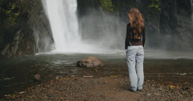 Güçlü Bali Şelalesi 'ne bakan turist kadın, taşlardan akan su. Uzun saçlar rüzgarda savruluyor. Turizm, yürüyüş, yürüyüş, rahatlama, aktif yaşam tarzı, yaz tatili. Yavaş arka plan