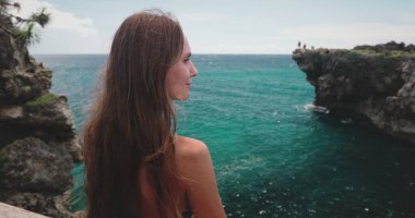 A woman stands confidently on the edge of a cliff overlooking the vast ocean on Wild Sumba Island in Indonesia. The Weekurri Lagoon Salt Lake sparkles in the distance. clipart