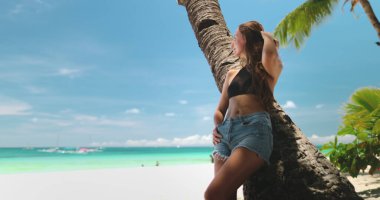 Genç kadın tropikal tatilinin tadını çıkarıyor. Beyaz bir kumsalda dinleniyor. Arka planda turkuaz su ve mavi gökyüzü olan bir palmiye ağacına yaslanıyor.