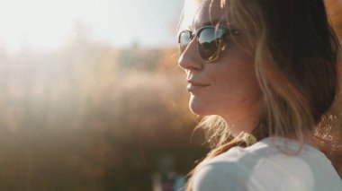 Retro güneş gözlüklü şık bir kadın gün batımına bakıyor. Sıcak, yumuşak ışıkta güzel bir kız portresi. Dişi yakın çekim kırsal orman manzarasının tadını çıkarıyor. Turuncu tonlama filtresi. İnsanlar dışarıda rahatlar. Yavaş çekim