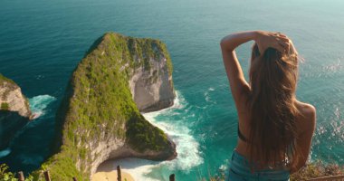 Genç turist kadın saçlarına dokunurken Kelingking 'in Nusa Penida, Bali' deki gizli sahili manzarasının tadını çıkarıyor.