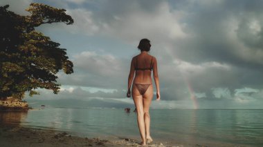 Bikinili bir kadın kumlu sahilde yürüyor, dalgalar arka planda yavaşça kıyıya vuruyor. Yaz tatilinde açık hava seyahati. Güzel doğa deniz manzarası