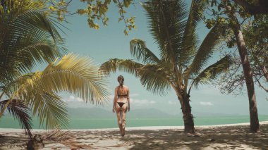 Genç bir kadın, arka planda turkuaz suyu olan palmiye ağaçlarıyla çevrili beyaz bir kumsalda yürürken tatilinin tadını çıkarıyor.