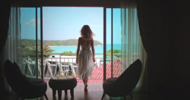 Uçan beyaz elbiseli bir kadın, Asya 'nın güneydoğusundaki turkuaz okyanusa bakan bir otel balkonuna doğru yürüyor. Huzurlu ve çarpıcı manzaranın tadını çıkarıyor.