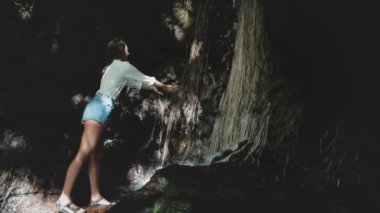 Turist kadın orman şelalesinin tadını çıkarıyor. Dişi, sıcak yağmur ormanlarında yüzünü kaynak suyuyla yıkar. Yürüyüş sonrası ferahlatıcı. Tayland açık hava seyahati. Dramatik koyu tonlama. Yavaş çekim