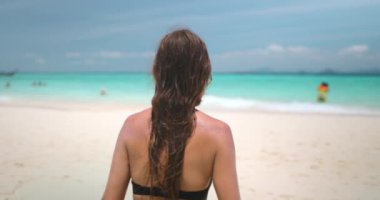 Bikinili bir kadın kumlu sahilde yürüyor, okyanus manzarası seyrediyor. Güneş gökyüzünde ışıl ışıl parlıyor. Turist kadın dinleniyor, yaz tatilinde açık havada seyahat ediyor. Arka plan, yavaş çekim