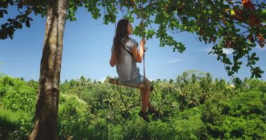 Tropikal orman bahçesindeki ağaçta sallanan kız. Uzun saçlı beyaz kadın yaz tatili tatilinde açık havada seyahat etmekten hoşlanıyor. Mavi gökyüzü ve yeşil yaprak ormanı. Yavaş çekim