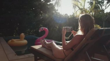 Bikinili bir kadın Tayland 'da havuz kenarında rahatlar, telefonla fotoğraf çeker, flamingo ve ördek yüzükleri yakınlarda yüzer. Kızım, yaz tatili tatili için özel villada dışarıda dinlen.