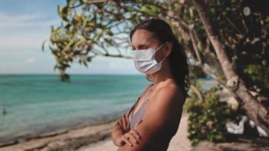 Yüz maskeli bir kadın sahilde tek başına duruyor, dalgalar arkasına vuruyor yaz güneşinin altında. Varlığı, dinginlik ve devam eden COVID-19 krizi arasındaki belirgin zıtlığı yansıtıyor..