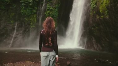 Güçlü bir Bali şelalesinin önünde bir kadın duruyor, su çağlayarak akıyor. Sahne doğanın güzelliğini ve gücünü yakalar..
