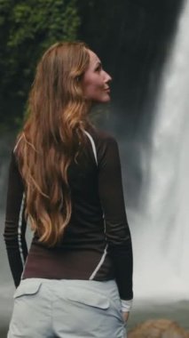 Dikey Ekran Videosu. Bereketli ormandaki görkemli şelalenin huzur verici güzelliği. Kadın huzur içinde duruyor. Rüzgarda uçuşan saçlar. Macera ve doğa bağlantısı için mükemmel bir yer. Bali 'ye Seyahat