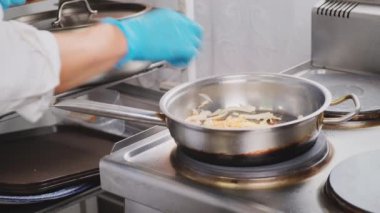 Şef soğan kızartıyordu. Çelik tepside kırmızı biberli. Kadın, elleri mavi eldivenle pişiriyor. Malzemeleri demir tavada karıştırıyor. Yakın çekim. Lokanta mutfağında yemek pişirmek, sağlıklı beslenme düzeni.