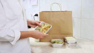 Lokantada yemek servisi. Şef beslenme çantalarını kese kağıdına koyar. Sağlıklı beslenme, günlük menü, plastik kaplarda yemek yemekleri. Kadın aşçı beyaz mutfakta yemek kutusu topluyor.