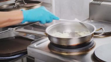 Mutfaktaki çelik tavada soğan kızartması yakın. Mutfak spatulasıyla kadın eli, hazır yemek için kesilmiş soğan malzemesini karıştırıyor. Lokantada aşçı, yemek pişirme, sağlıklı beslenme.