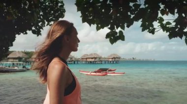 Turist kız Bora Bora su villasının deniz manzaralı köşkünün tadını çıkarıyor. Beyaz kadın ağaçların arkasındaki manzaranın altında sahilde duruyor. Yaz tatilinde açık havada hayat yolculuğu, Fransız Polinezyası