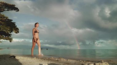 Gökkuşağının altında bikinili bir kadın tropikal sahilde yürüyor. Dramatik akşam gün batımı fırtınası üstüne yağmur. Yaz tatilinde açık hava seyahati. Güzel doğa deniz manzarası