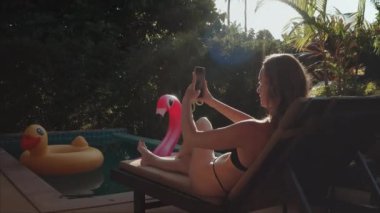 Genç bir kadın havuz kenarında güneşlenirken akıllı telefonuyla selfie çekiyor. Güneş parlıyor ve Tayland 'da tatilinin tadını çıkarıyor.