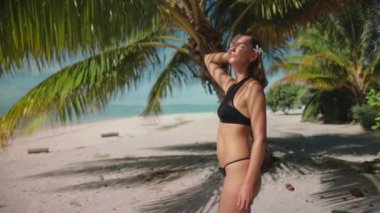 Bikinili bir kadın kumlu plajda duruyor ve arka planda dalgalar çarpıyor. Güneş tepemizde ışıldıyor.