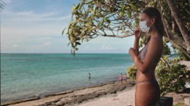Plajdaki kadın tıbbi maskeyi çıkar ve normal hayatın tadını çıkar. Tropikal adada yaz tatili. Karantina ve kendini izole etme, salgın salgın hastalık, kovid koronavirüs sonu, konsept karantina.