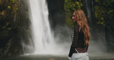 Yeşilliklerle çevrili görkemli bir Bali şelalesinin önünde bir kadın duruyor. Su taşar ve nefes kesen doğal bir manzara yaratır..