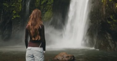 Güçlü bir orman şelalesinin yanında duran turist kadın. Uzun saçlar rüzgarda savruluyor. Turizm, yürüyüş, yürüyüş, rahatlama, aktif yaşam tarzı, Bali 'de yaz tatili. Yavaş çekim 4K arka görünüm yakınlaştır