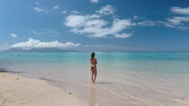 Bikini giyen bir kadın kumlu sahilde duruyor, arkasında okyanus dalgaları var. Mavi gökyüzü güneşli bir gün. Tahiti, Fransız Polinezyası