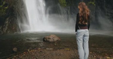 Güçlü Bali Şelalesi 'ne bakan turist kadın, taşlardan akan su. Uzun saçlar rüzgarda savruluyor. Turizm, yürüyüş, yürüyüş, rahatlama, aktif yaşam tarzı, yaz tatili. Yavaş çekim geri görünümü