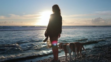 Kadın ve köpekleri sakin bir plajda gün batımının keyfini çıkarıyorlar. Doğada sevgi ve arkadaşlık manzarası yaratıyorlar.