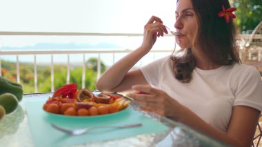 Güzel bir kadın, tropikal manzaraya tepeden bakan bir balkonda otururken egzotik meyvelerin tadını çıkarıyor.