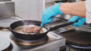 Profesyonel bir restoran mutfağında aşçı sıcak tavada eti karıştırır ve çevirir. Mavi eldivenli profesyonel aşçı demir tavada tavuk ciğeri kızartıyor. Yemek yapmak, müşteriler için malzeme hazırlamak.