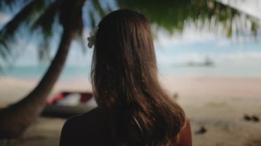 Genç bir kadın tropik bir plajda, bir palmiye ağacının altında, saçında geleneksel bir çiçekle rahatlatıcı bir anın tadını çıkarıyor. Hafif deniz meltemi hafifçe saçlarını savuruyor.