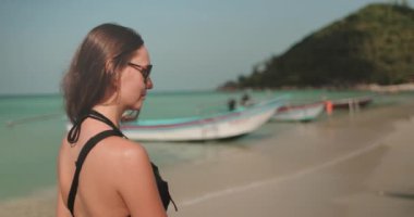 Güneş gözlüklü bir kadın kumlu sahilde yürüyor, sığ sularda uzun kuyruklu tekneler. Arka planda tropik plaj ve berrak mavi su. Turizm, tatil, egzotik Tayland adasına seyahat. Yavaş çekim 4K