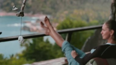 Bir kadın, balkonun kenarına kurulmuş bir sandalyede oturuyor. Manzaraya bakıyor. Rahatlamış ve dalgın görünüyor. Etrafı inceliyor..