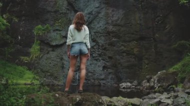 Genç turist, Moorea 'da güneşli bir günde yemyeşil bir tropikal ormandaki şelale manzarasının tadını çıkarıyor. Fransız Polinezyası.