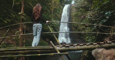 Tourist woman travel Bali island explore Melanting Waterfalls. Long hair hiker girl walk bamboo bridge enjoy view on powerful waterfall in jungle tropical forest. Outdoor holiday vacation. Slow clipart