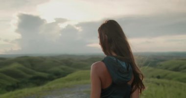 Genç kadın Endonezya 'da bir tepede duruyor, yemyeşil tepelerin üzerinde gün batımını izliyor, turuncu ve pembe renklerle gökyüzünü boyuyor.