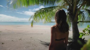Tropik bir sahilde palmiye ağacının altında duran bir kadın manzaranın tadını çıkarıyor. Güneş parlıyor, dalgalar çarpıyor, huzurlu bir rahatlama ve mutluluk sahnesi yaratıyor.
