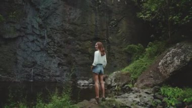 Tropikal bir ormanda kayalık bir uçurumdan akan şelaleye hayran bir turist. Bereketli bitki örtüsüyle çevrili bir huzur ve sükunetin tadını çıkarıyor.