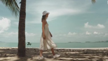 Beyaz elbiseli ve şapkalı zarif bir kadın Tayland 'da kumsalda yürüyor. Palmiye ağaçları, mavi gökyüzü, sakin okyanus. Turist kız dinleniyor, deniz manzarasının tadını çıkarıyor. Yaz tatilinde seyahat