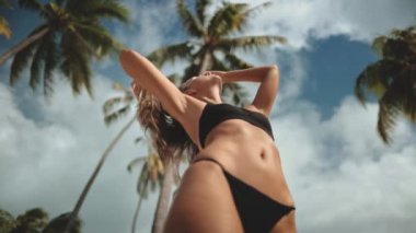 Genç bir kadın tropik bir plajda dikiliyor, palmiye ağaçlarıyla çevriliyken güneşin ve esintinin tadını çıkarıyor. Sıcak güneş ışığı ve yumuşak rüzgar huzur ve sükunet hissi yaratır.