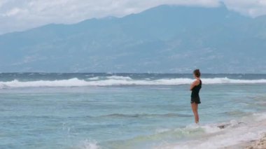 Genç bir kadın okyanusta yürüyor, kıyıya vuran dalgaların tadını çıkarıyor ve uzaktaki dağları seyrediyor.