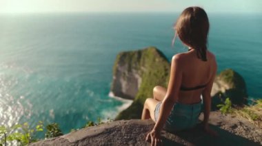 Kadın, Endonezya 'nın Nusa Penida Adası' ndaki okyanus manzarasına bakan bir uçurumun kenarında oturuyor. Seyahat, turizm, tatil, tatil, yürüyüş. Ağır çekim. Arka plan