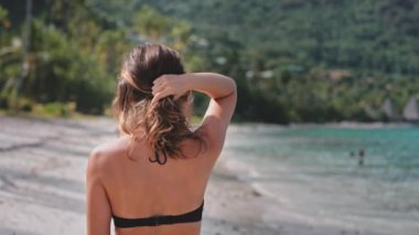 Kumsalın kumlu sahilinde yürüyen siyah bikinili bir kadın. Güneş parlıyor ve dalgalar yavaşça kıyıya vuruyor..